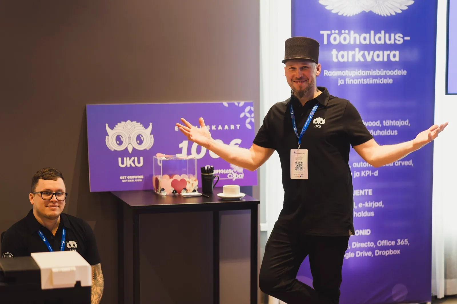 Uku boks Meriti Inspiratsioonipäeval 2026: Uku tiimi liige seisab käed laiali tööhaldustarkvara bänneri ees, teine istub laua taga, taustal Uku 100 euro elamuskaardi plakat